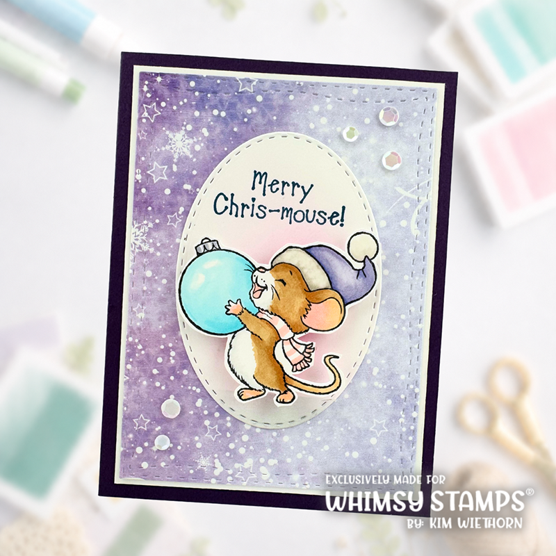 A handmade greeting card featuring a cute mouse character in a blue and white hat hugging a light blue ornament, with the text 'Merry Chris-mouse!' on a purple watercolor background. The card is displayed on a light surface with craft supplies blurred in the background.