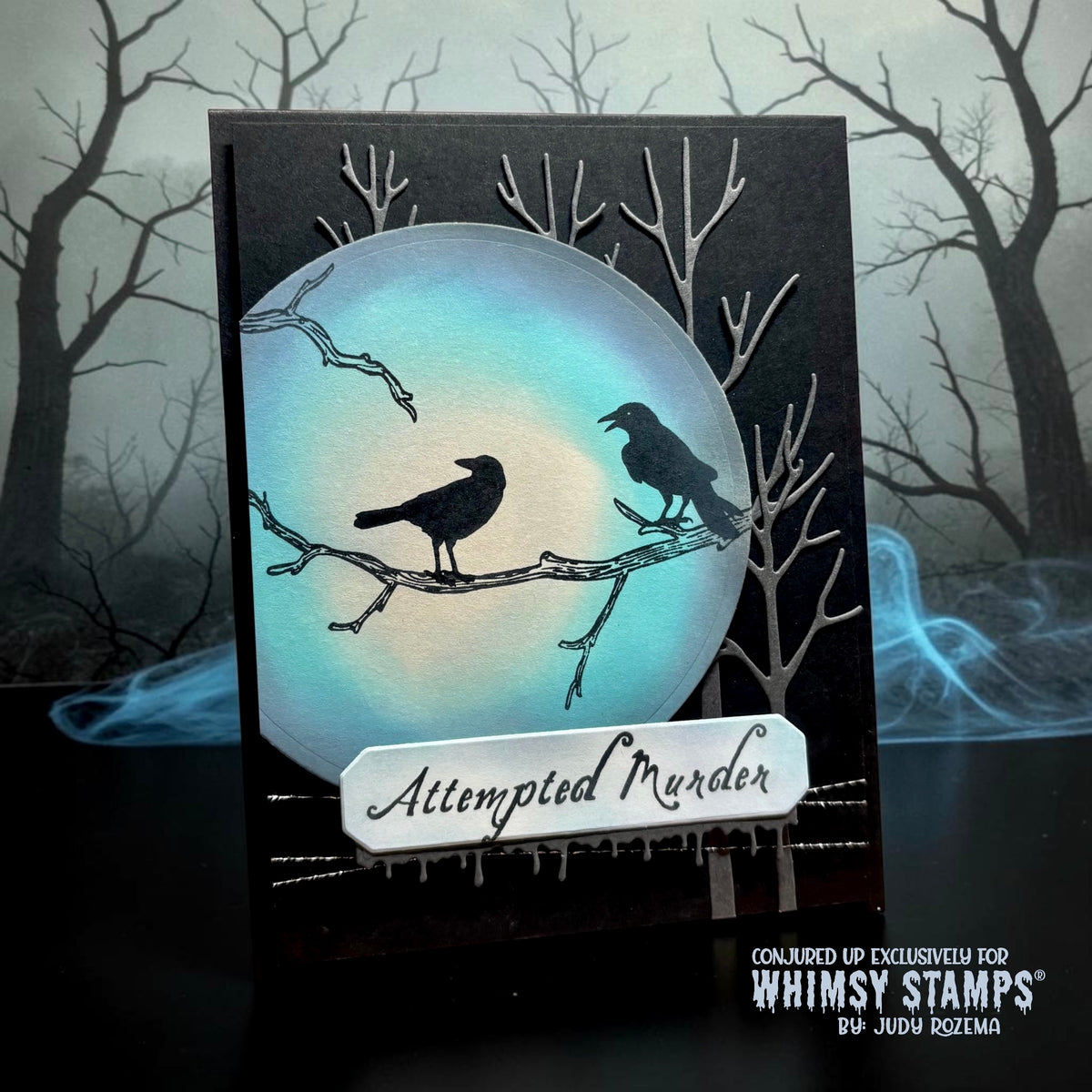 A handmade gothic-style card featuring two black crows perched on bare tree branches in front of a glowing blue-white full moon. There is a rectangular strip that reads 'Attempted Murder.'